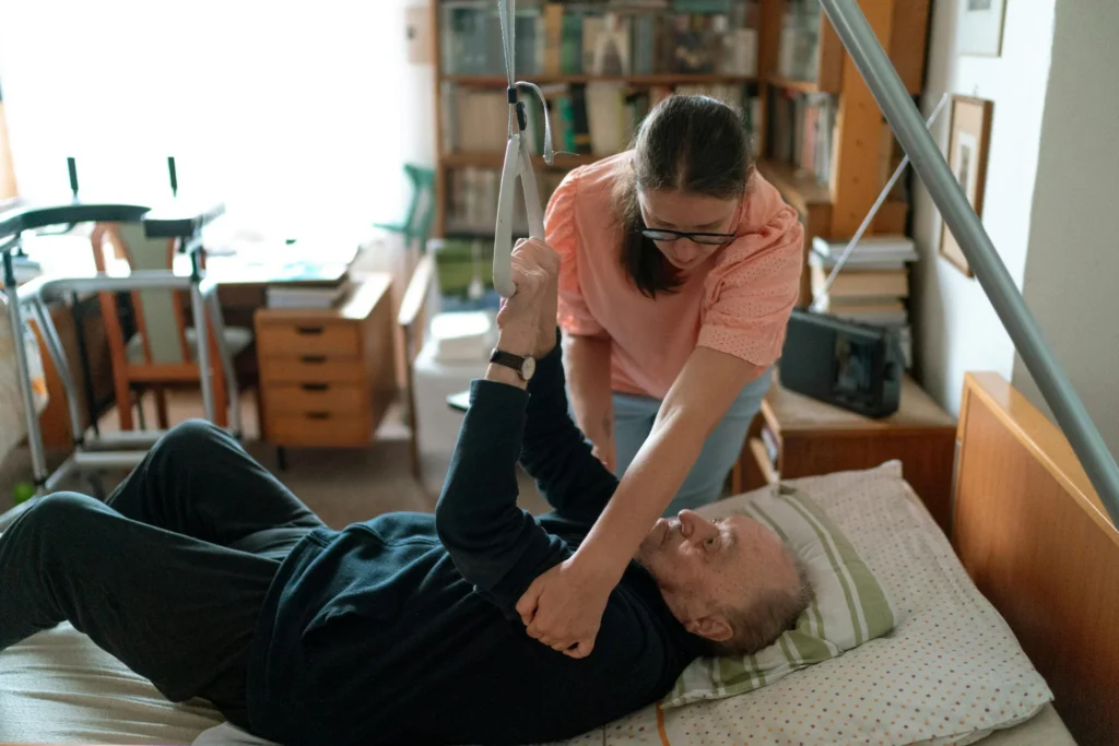 Opiekunka pomaga seniorowi zmienić pozycję w łóżku rehabilitacyjnym, senior przytrzymuje się uchwytu wysięgnika.